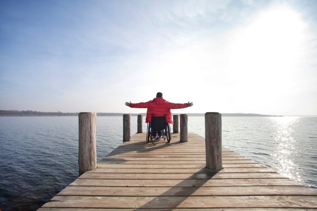 A backside view of a person in a wheelchair on a dock overlooking water. Their hands are spread wide. It is implied that they're a Palco participant.
