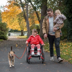 A medium close-up view of a father out walking the family dog with his young daughter who is a wheelchair user he is carrying his youngest in his arms and his older daughter is holding the pet leash as they enjoy the day together.