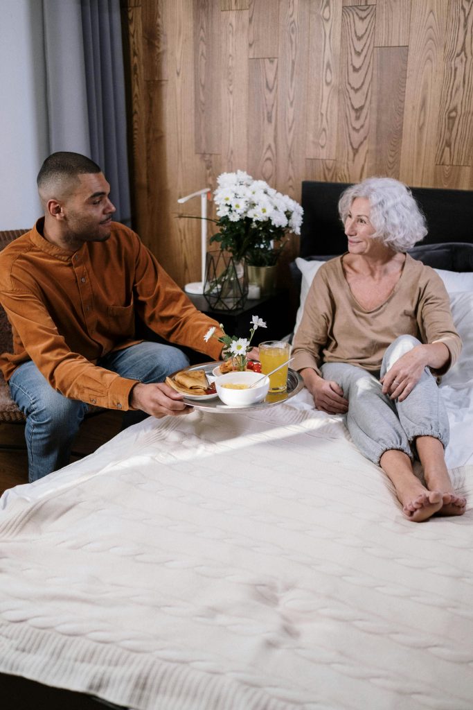 A woman with wavy white hair cut into a bob is seated in bed. She is wearing comfortable clothing and smiling toward the man seated next to her, holding out a tray to her, filled with a nutritious breakfast. She uses self-direction