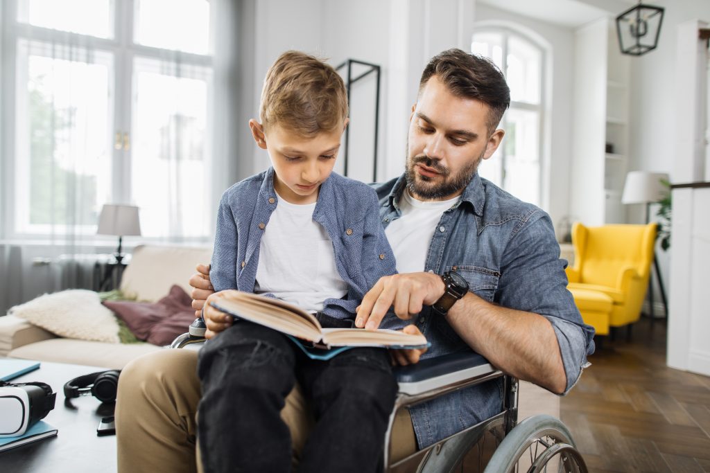 A dad in a wheelchair is reading a book with his son, who is sitting on his lap, in their home.