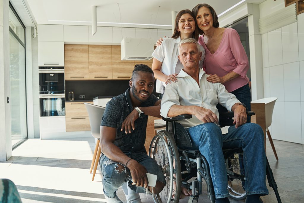 A white man in a wheelchair smiles at the camera while surrounded by three of his caregivers, who are also smiling.